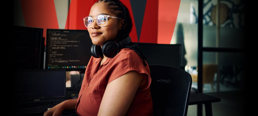 Smiling black woman with headphones around her neck sitting in front of 3 monitors