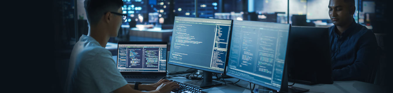 Young man in glasses working in front of 3 monitors
