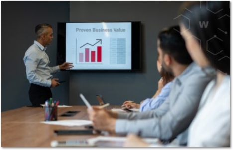 Person presenting a chart with red bars and an arrow on top in front of a table of co-workers